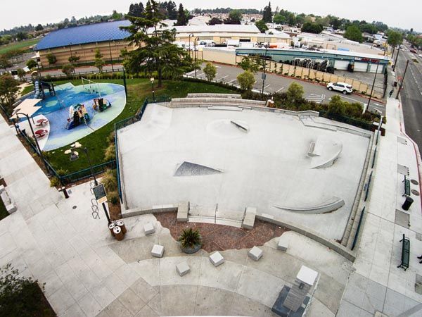 Aerial view of a white skatepark beside a colorful splash pad and surrounding buildings.