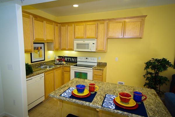 Compact kitchen with light wood cabinets, white appliances, and a table set with colorful place settings.