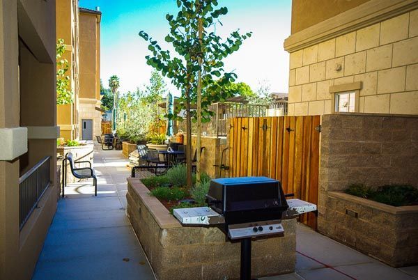 Apartment courtyard with grill, wooden fence, potted plants, and a walkway in sunlight