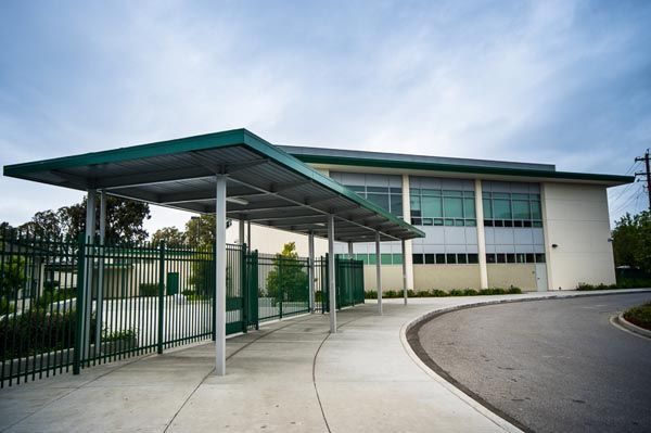 Modern building entrance with green metal canopy, fenced walkway, and curved driveway under a cloudy sky
