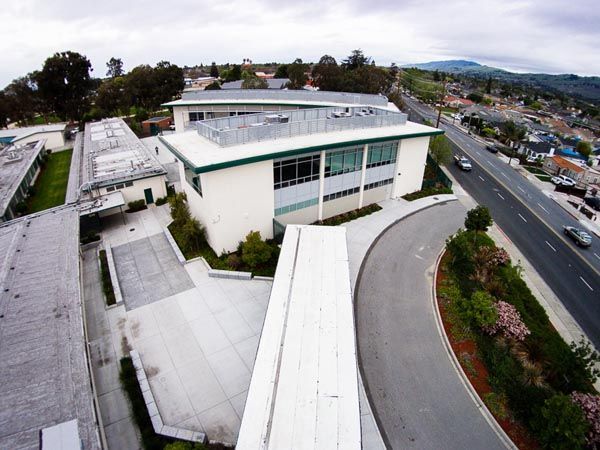 Aerial view of a modern white building beside a road, with hills and a suburban neighborhood in the background