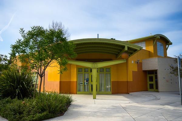 Modern yellow building with curved roof, glass doors, and landscaped entrance under a blue sky