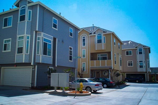 Apartment complex with blue, tan, and gray buildings, parked cars, and a sunny courtyard driveway