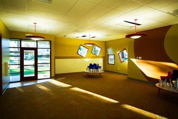 Empty yellow lobby with sunlight, glass doors, tilted wall frames, and a small table of chairs.