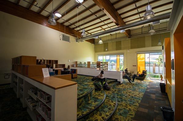 Open library with bookshelves, tables, and colorful carpet under a high wood-beam ceiling.