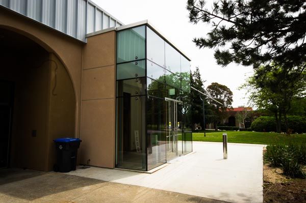 Modern glass entrance beside a tan building, opening onto a bright walkway and green lawn