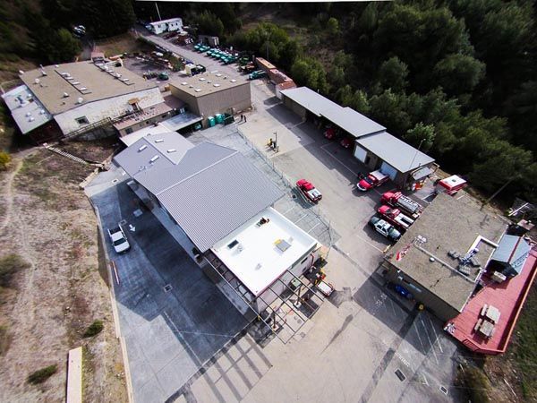 Aerial view of a parking lot with several cars and low buildings surrounded by trees.