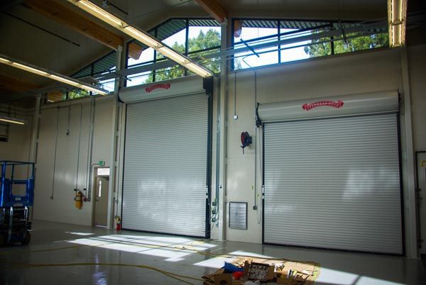 Industrial garage with two closed white roll-up doors and overhead lights inside a warehouse.