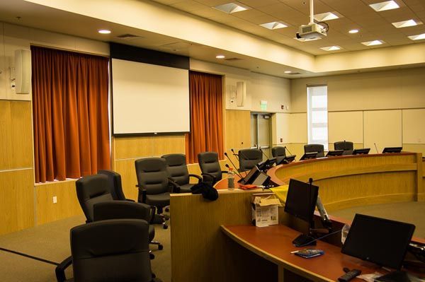 Empty conference room with desks, monitors, black chairs, and a projector screen behind red curtains.