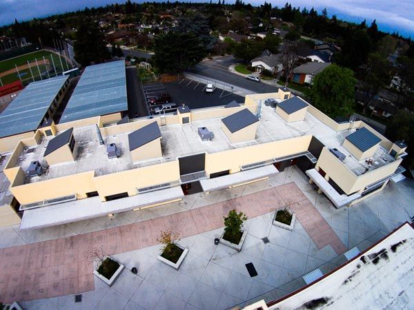 Aerial view of a beige building with rooftop skylights beside a paved courtyard and trees.