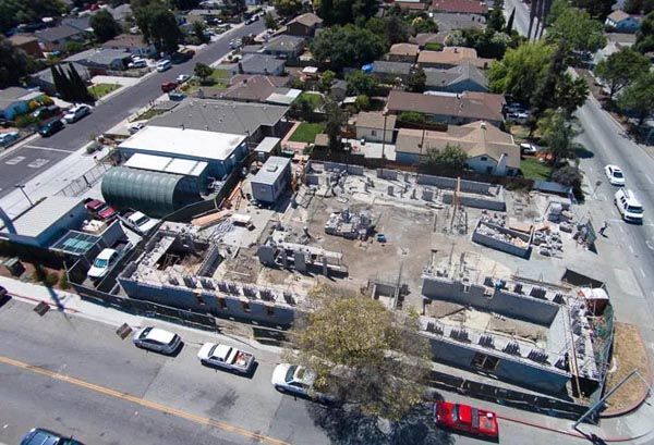 Aerial view of a construction site beside a street, with houses and parked cars nearby.
