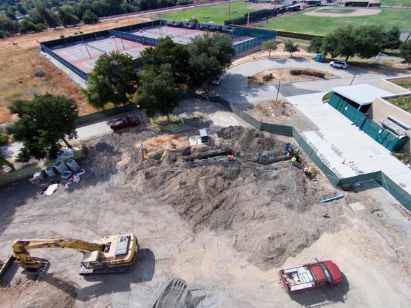 Aerial view of a construction site with excavators, trucks, and a large dirt excavation near tennis courts.