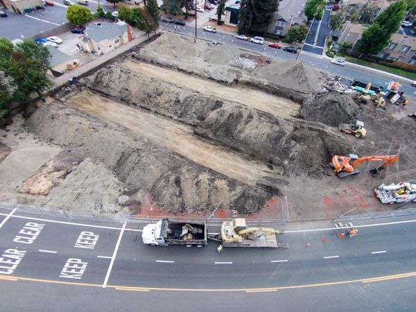 Aerial view of a road construction site beside a street, with excavators and trucks in a large dug-out area.