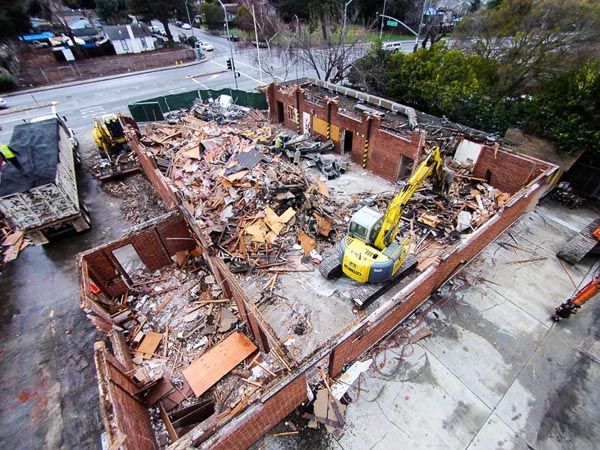 Collapsed brick building with debris and a yellow excavator at a demolition site