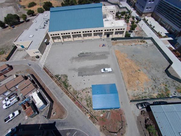 Aerial view of a large blue-roofed building beside an open paved lot and dirt field.
