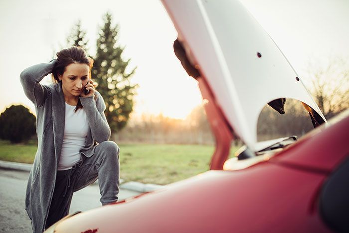 Woman kneeling by a red car with the hood open, looking concerned at sunset.