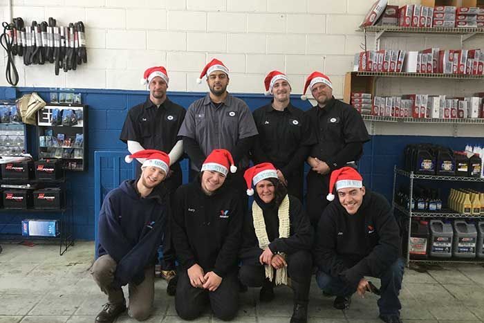Eight people wearing Santa hats pose in a workshop or garage with shelves and tools behind them.