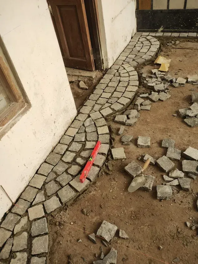 Sendero de piedra parcialmente terminado junto a un edificio, con adoquines sueltos y escombros esparcidos por el suelo.