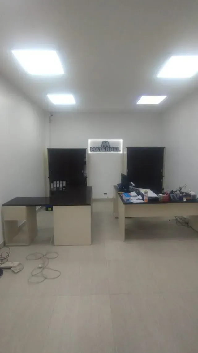 Oficina pequeña con paredes blancas, dos escritorios, monitores negros y luces en el techo.
