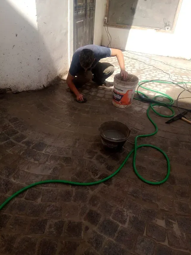Una persona arrodillada en un patio mojado, limpiando con un cubo y una manguera verde junto a una pared blanca
