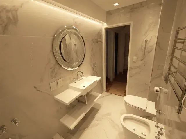 Baño moderno de mármol con espejo redondo, lavabo suspendido y váter junto a una puerta abierta