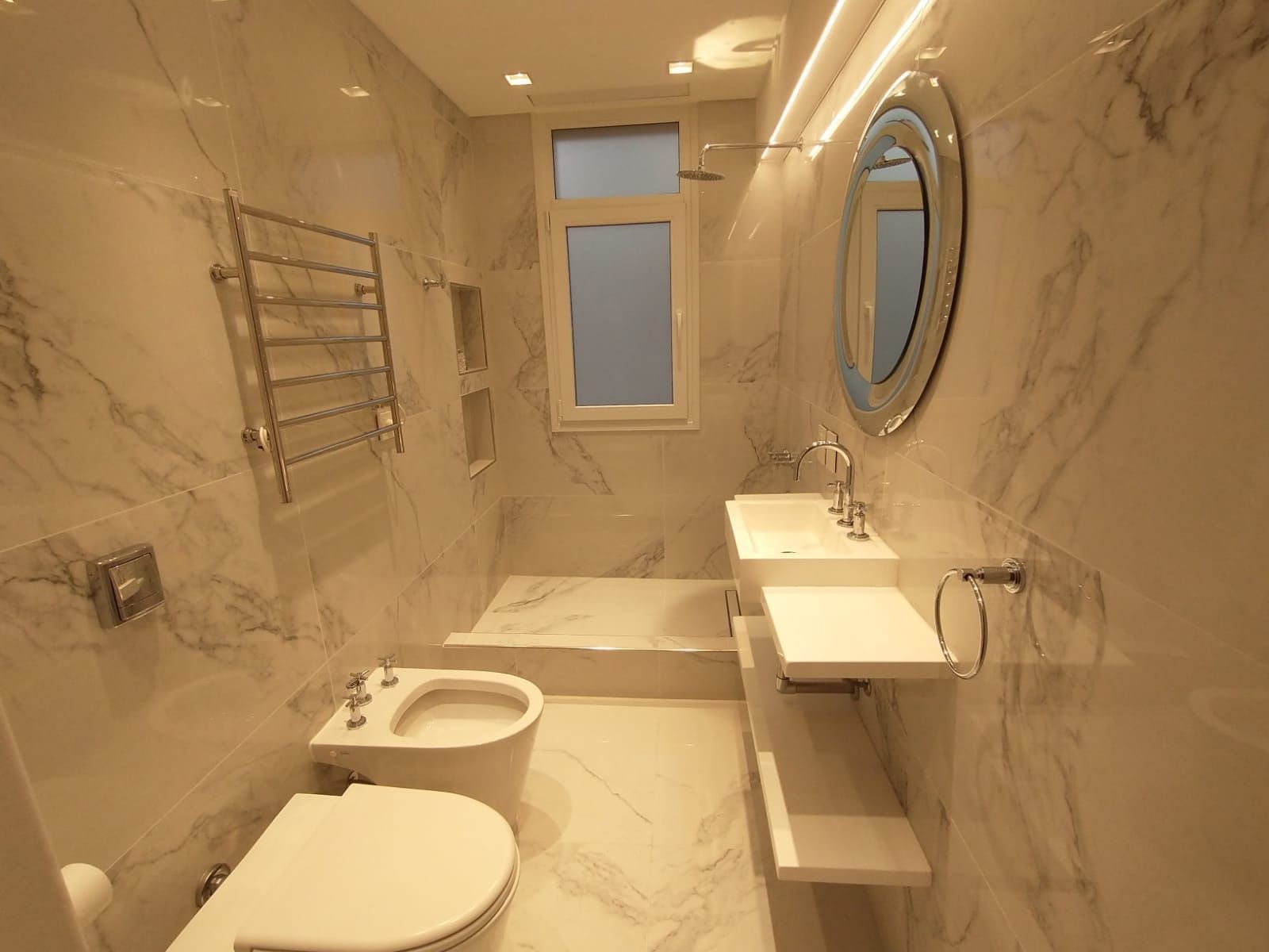 Baño moderno con azulejos de mármol, inodoro, lavabo, bañera, toallero y espejo redondo