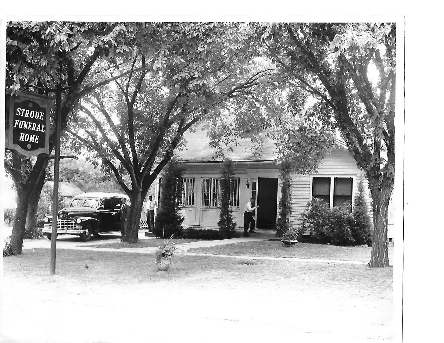 Black-and-white photo of a small house with trees, a parked car, and a sign in the yard