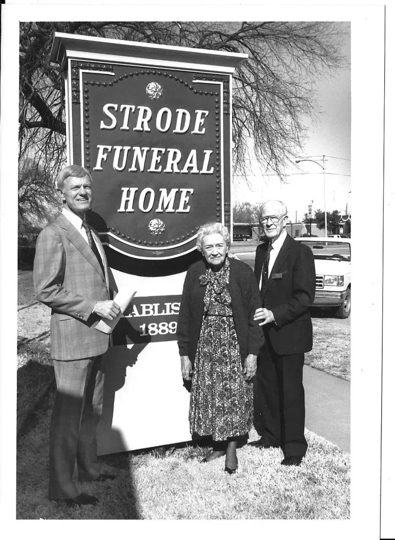 Three people standing beside a “Strode Funeral Home” sign outdoors in winter.