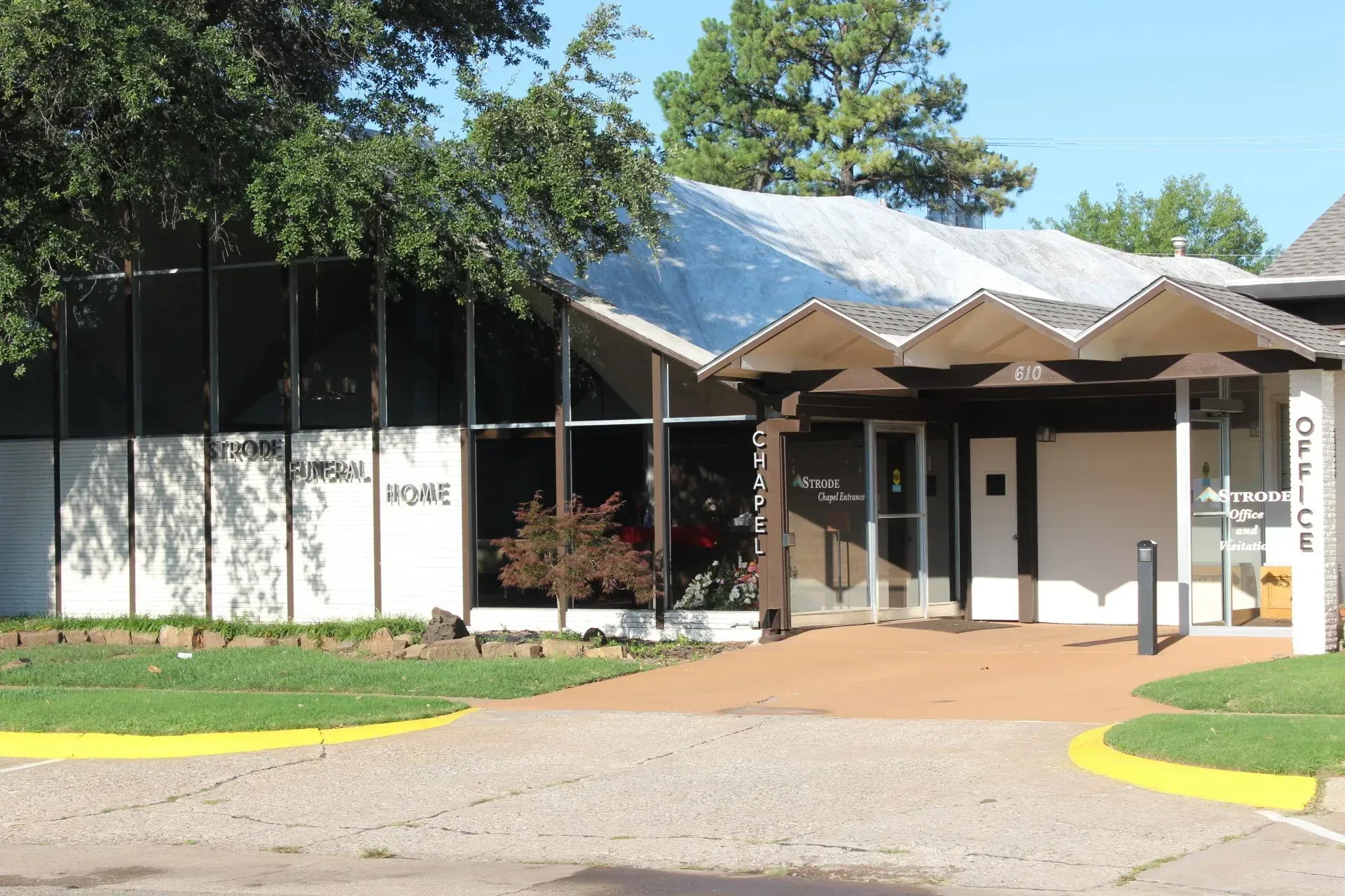Modern building entrance with glass walls, tan awning, and brick walkway under trees
