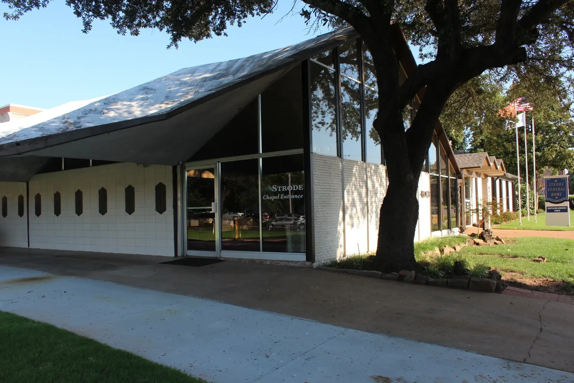 Modern white building with slanted roof and large windows beside a tree-lined sidewalk
