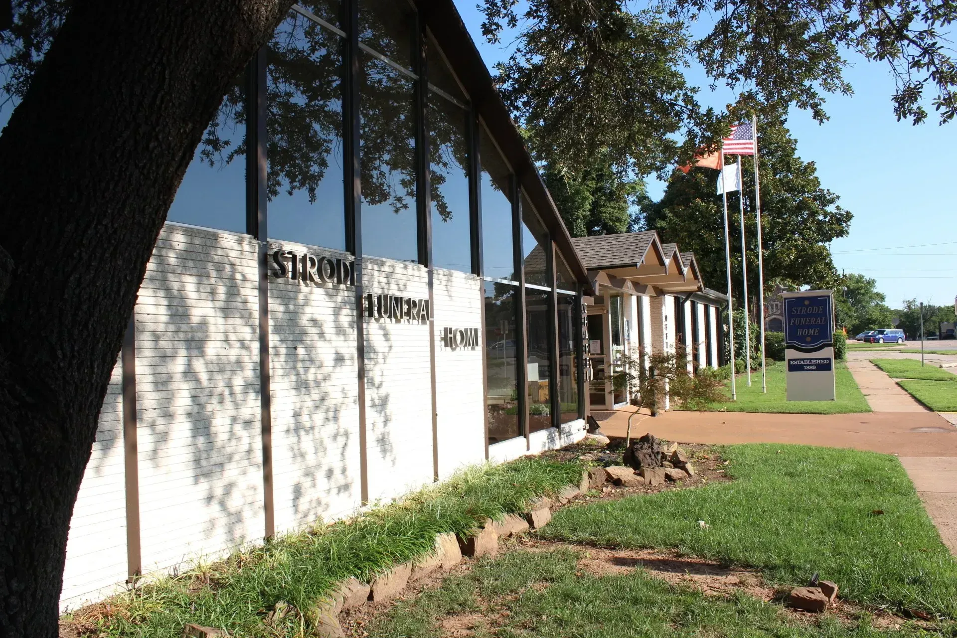 Building with white wall and glass windows beside a sidewalk, with a flagpole and grassy lawn on a sunny day