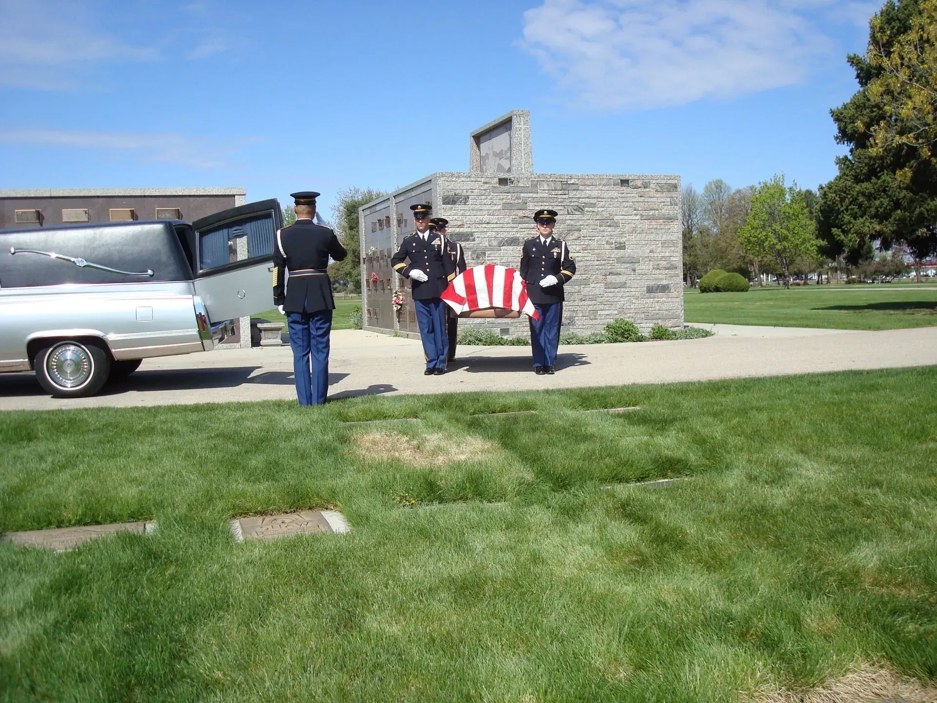Three uniformed officers stand by a casket draped with an American flag beside a hearse.