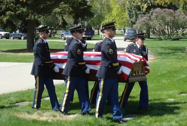 Four uniformed military pallbearers carry a flag-draped casket across a grassy lawn.