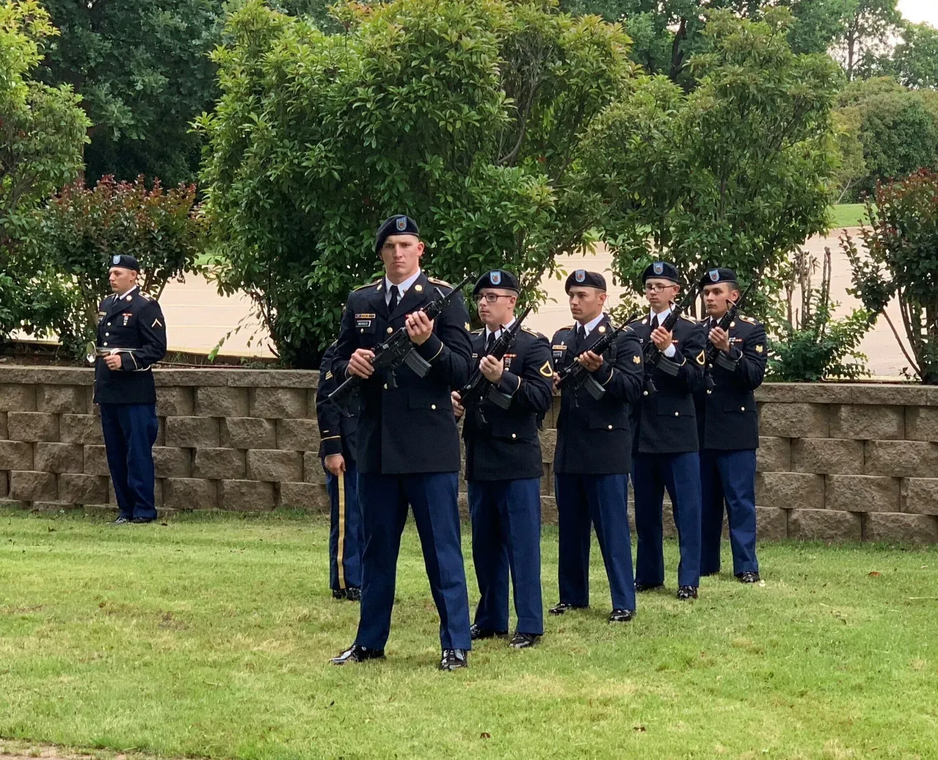 Uniformed officers standing in formation outdoors on grass, clapping in front of a stone wall and trees