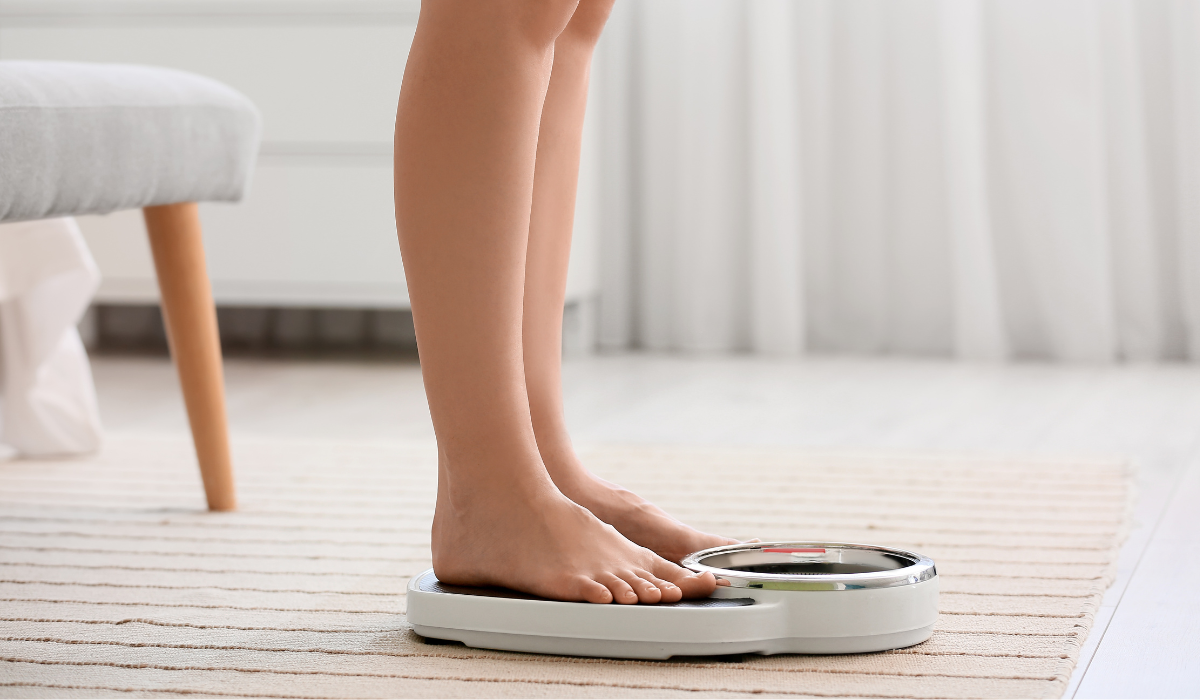 Person standing on a digital bathroom scale on a light rug near a bed and curtain