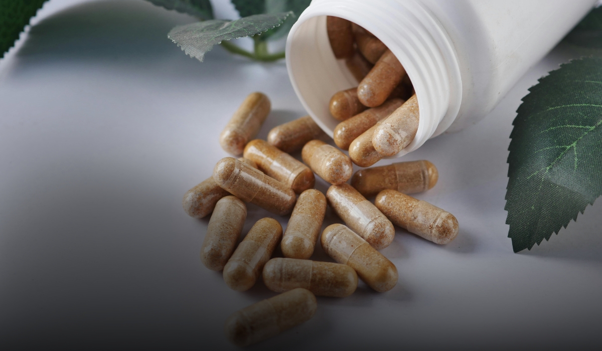 White pill bottle spilling brown capsules onto a gray surface with green leaves nearby