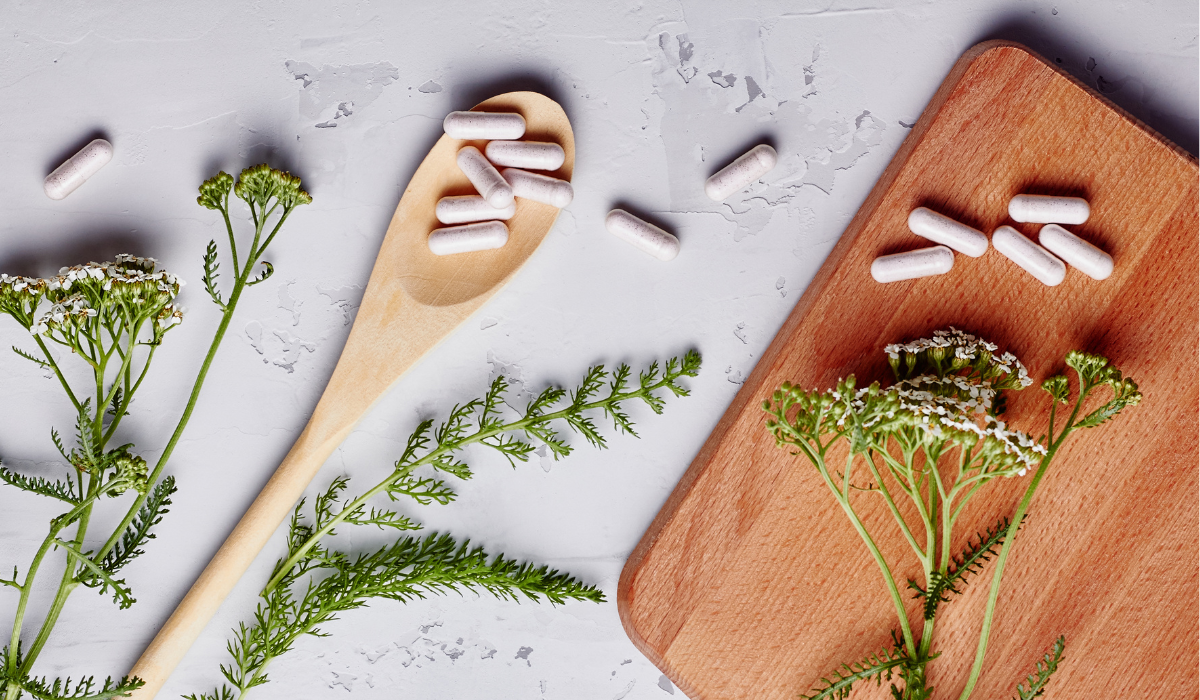 Assorted supplement capsules spilled onto a wooden spoon and cutting board, with a few surrounding herbal plants