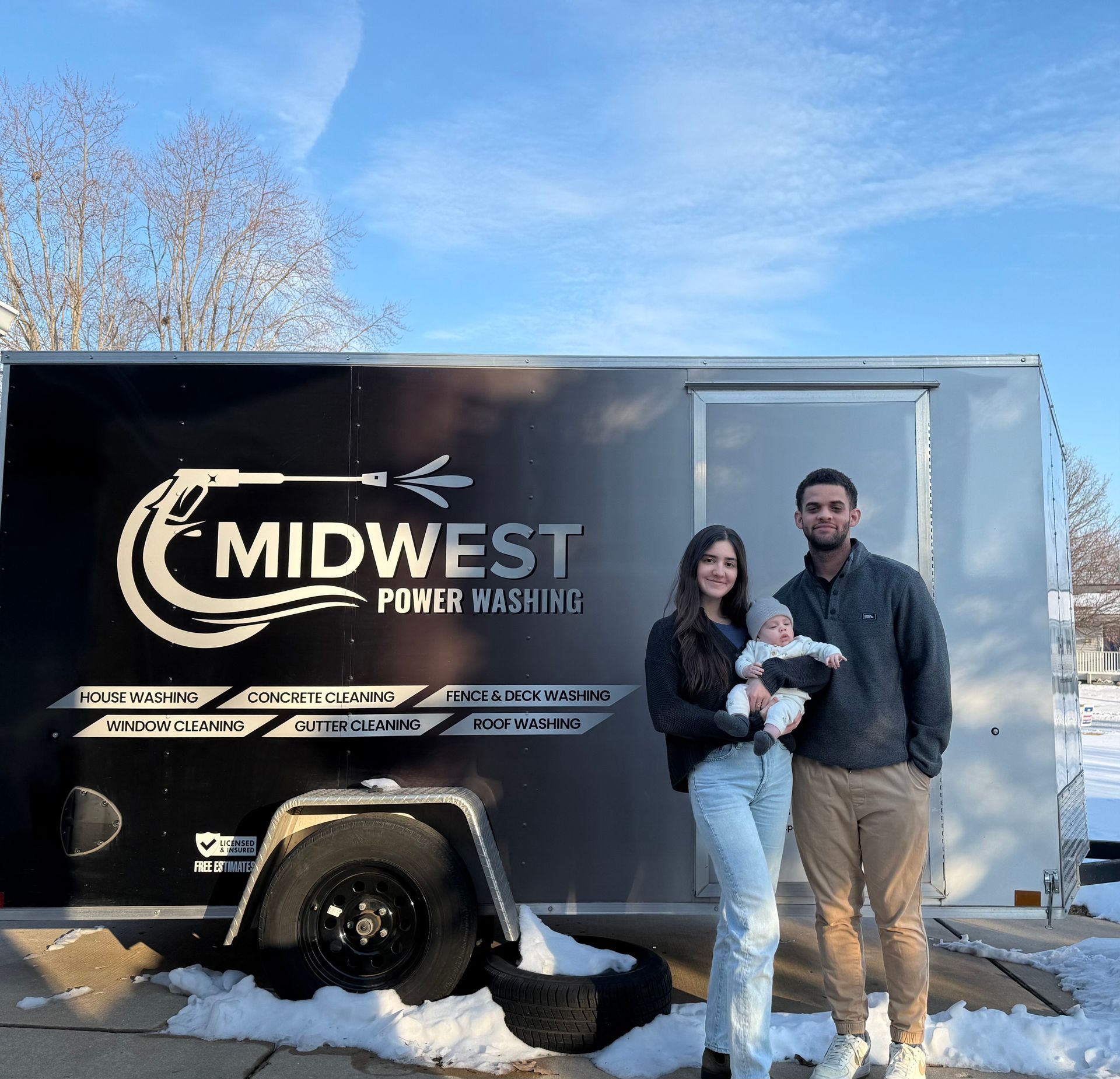 Family standing in front of a trailer for a power washing business. Baby is being held, snowy ground. Family standing in front of a trailer for a power washing business. Baby is being held, snowy ground.