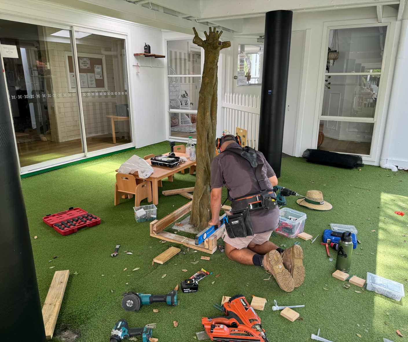 Person kneels on green carpet, assembling a wooden structure amid scattered tools in a bright room.