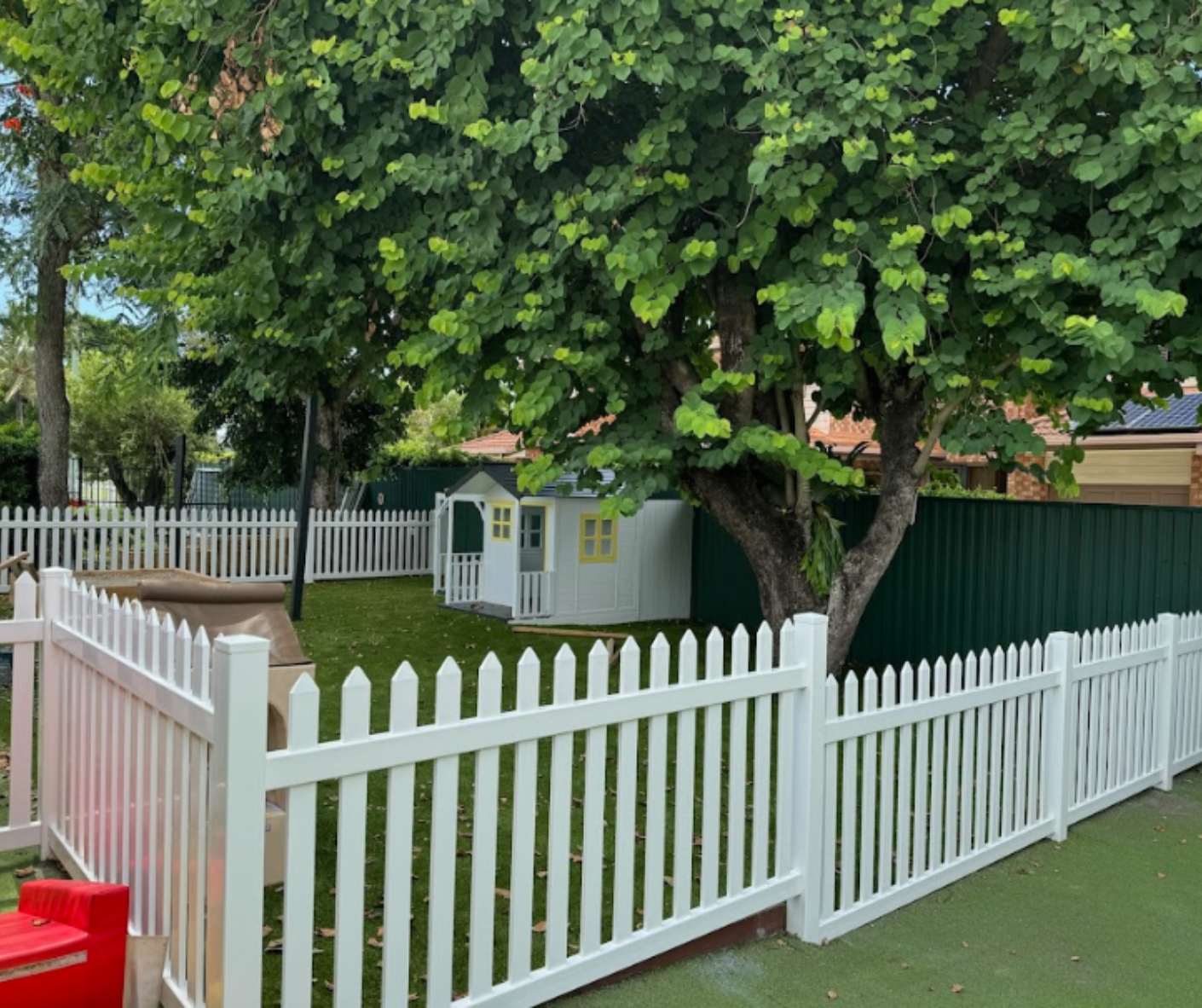 White picket fence around a small yard with a large leafy tree and a tiny house behind it