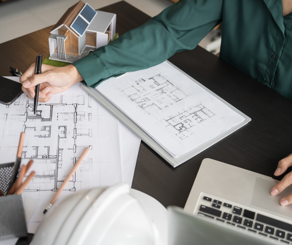Person reviewing house plans with a pencil, laptop, and architectural model on a desk