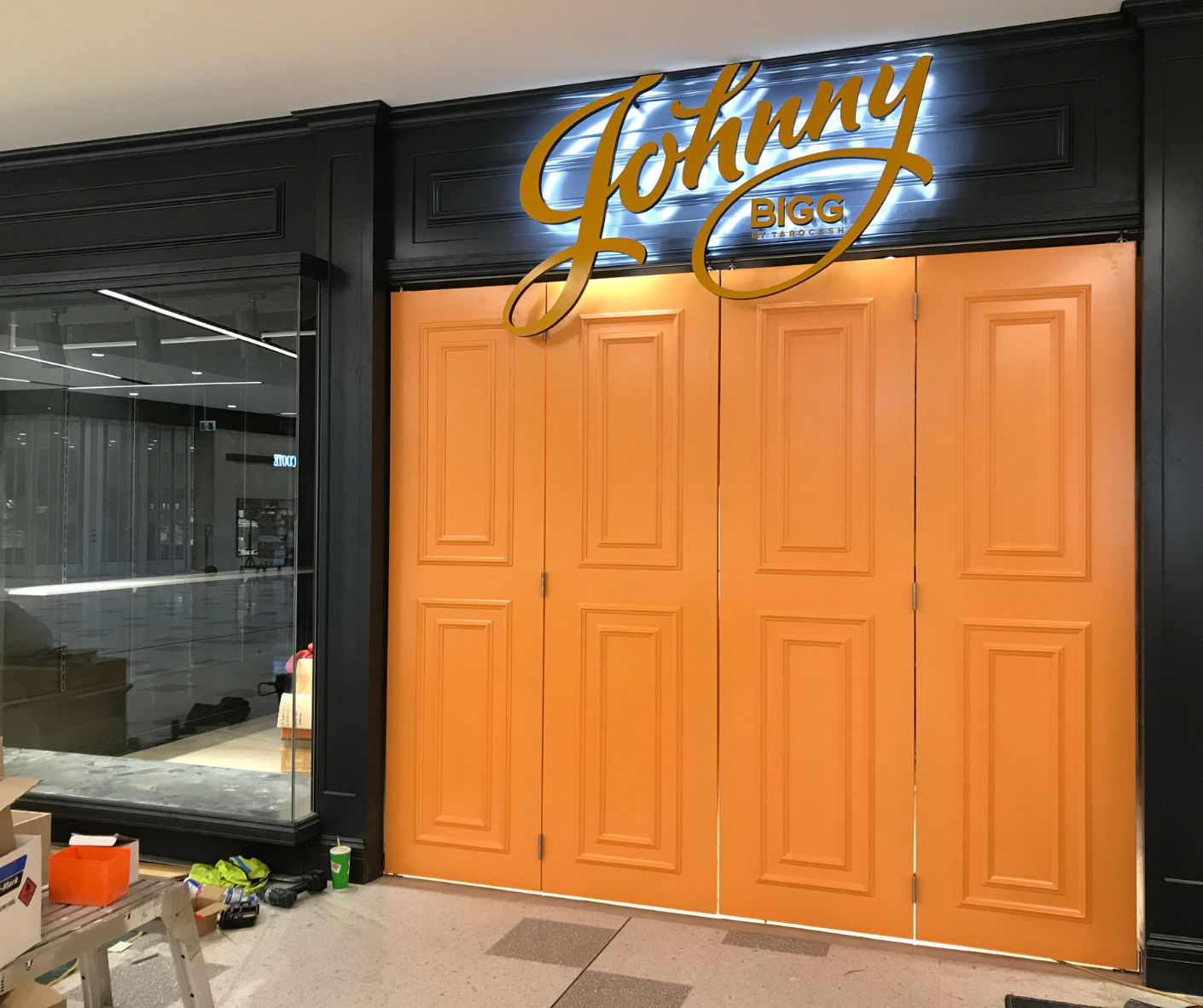 Orange double doors beneath a glowing “Johnny” sign in a modern storefront corridor Orange double doors beneath a glowing “Johnny” sign in a modern storefront corridor