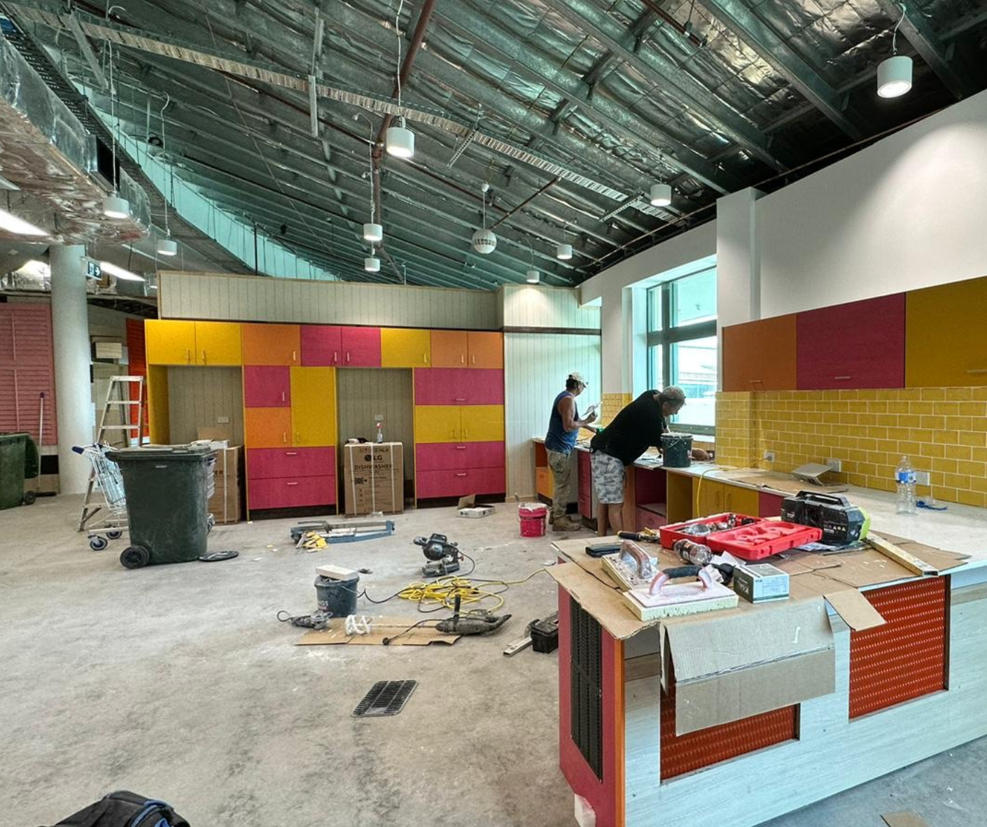 Renovation in progress inside a large room with exposed ceiling, colorful cabinets, tools, and workers near a counter Renovation in progress inside a large room with exposed ceiling, colorful cabinets, tools, and workers near a counter