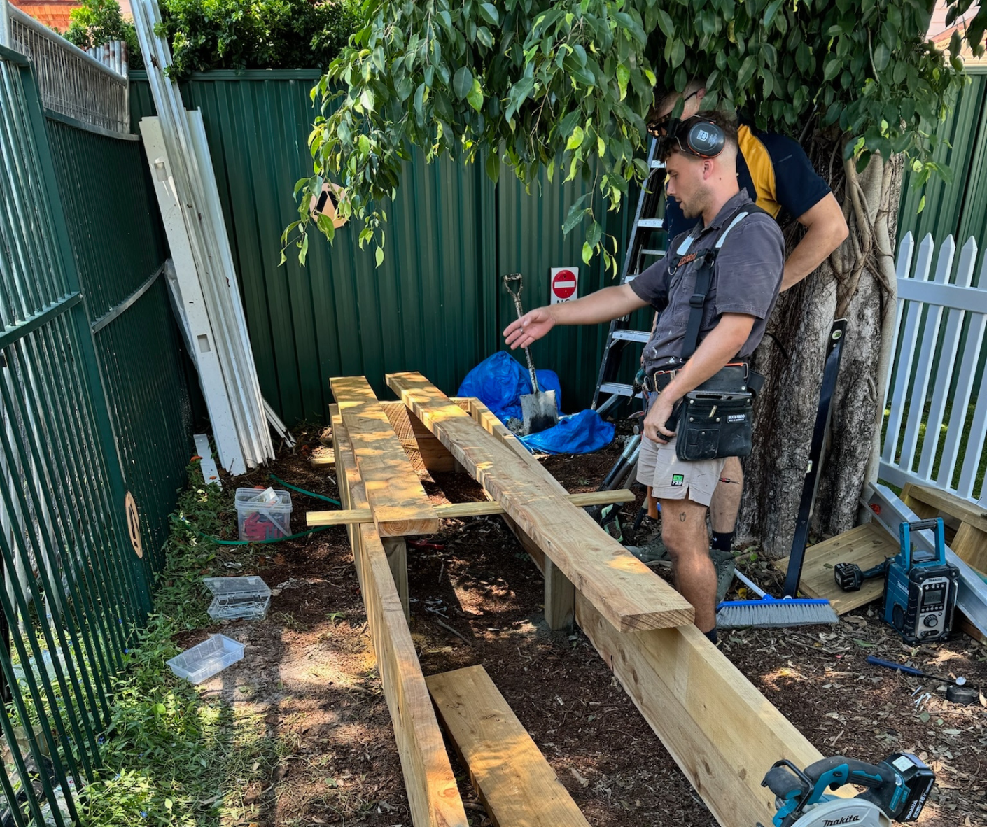 Two workers building a wooden deck beside a tree and green fence, with lumber and tools on the ground. Two workers building a wooden deck beside a tree and green fence, with lumber and tools on the ground.