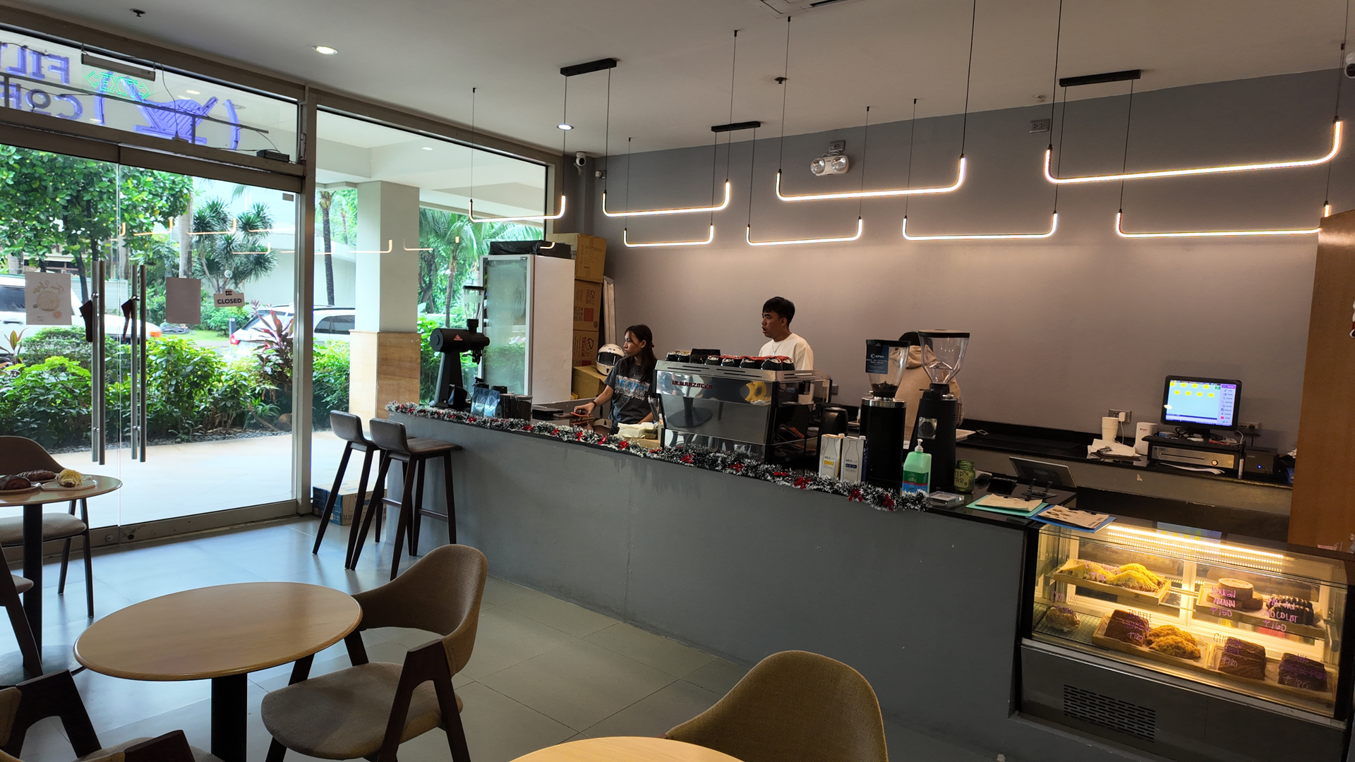 Small café counter with espresso machine, barista service area, geometric pendant lights, and a refrigerated cake display beside the front entrance. Small café counter with espresso machine, barista service area, geometric pendant lights, and a refrigerated cake display beside the front entrance.