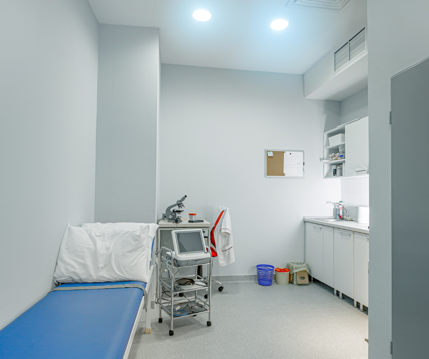Small medical exam room with a blue exam bed, rolling cart, desk, and white cabinets. Small medical exam room with a blue exam bed, rolling cart, desk, and white cabinets.