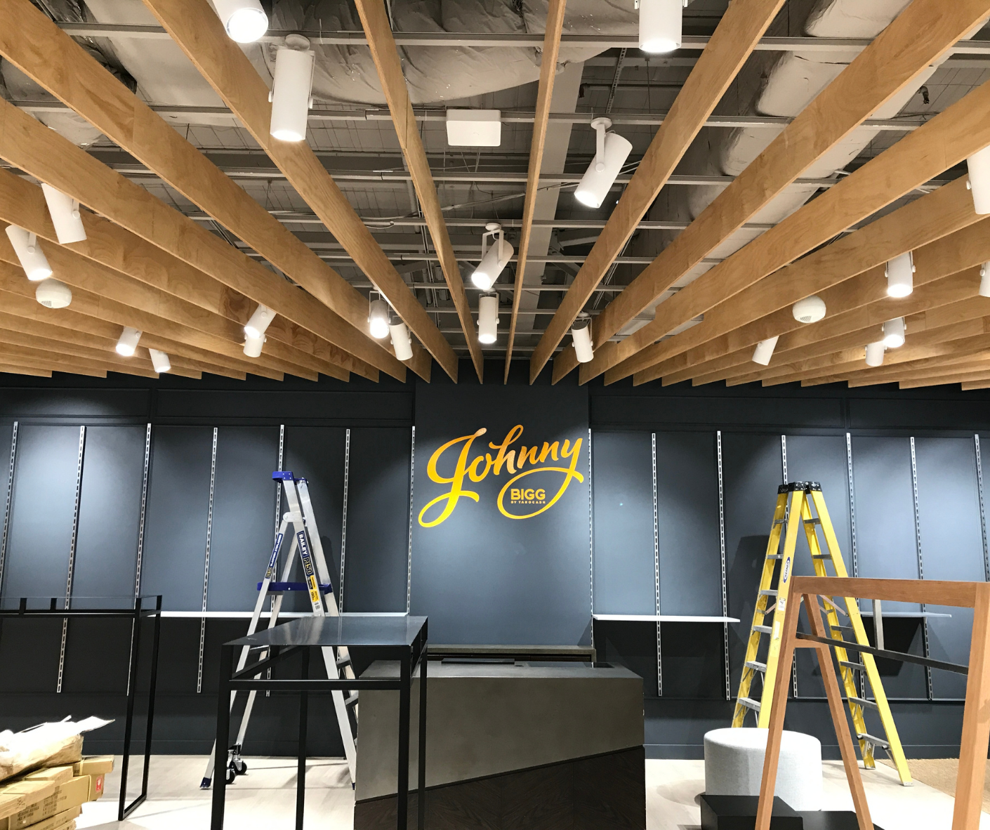 Indoor stage setup with wooden ceiling beams, blue backdrop, ladders, and a yellow “Johnny” logo center stage