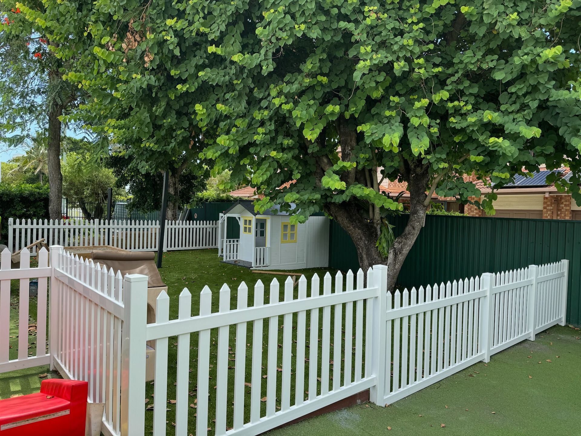 A white picket fence surrounds a backyard with a playhouse and a tree.