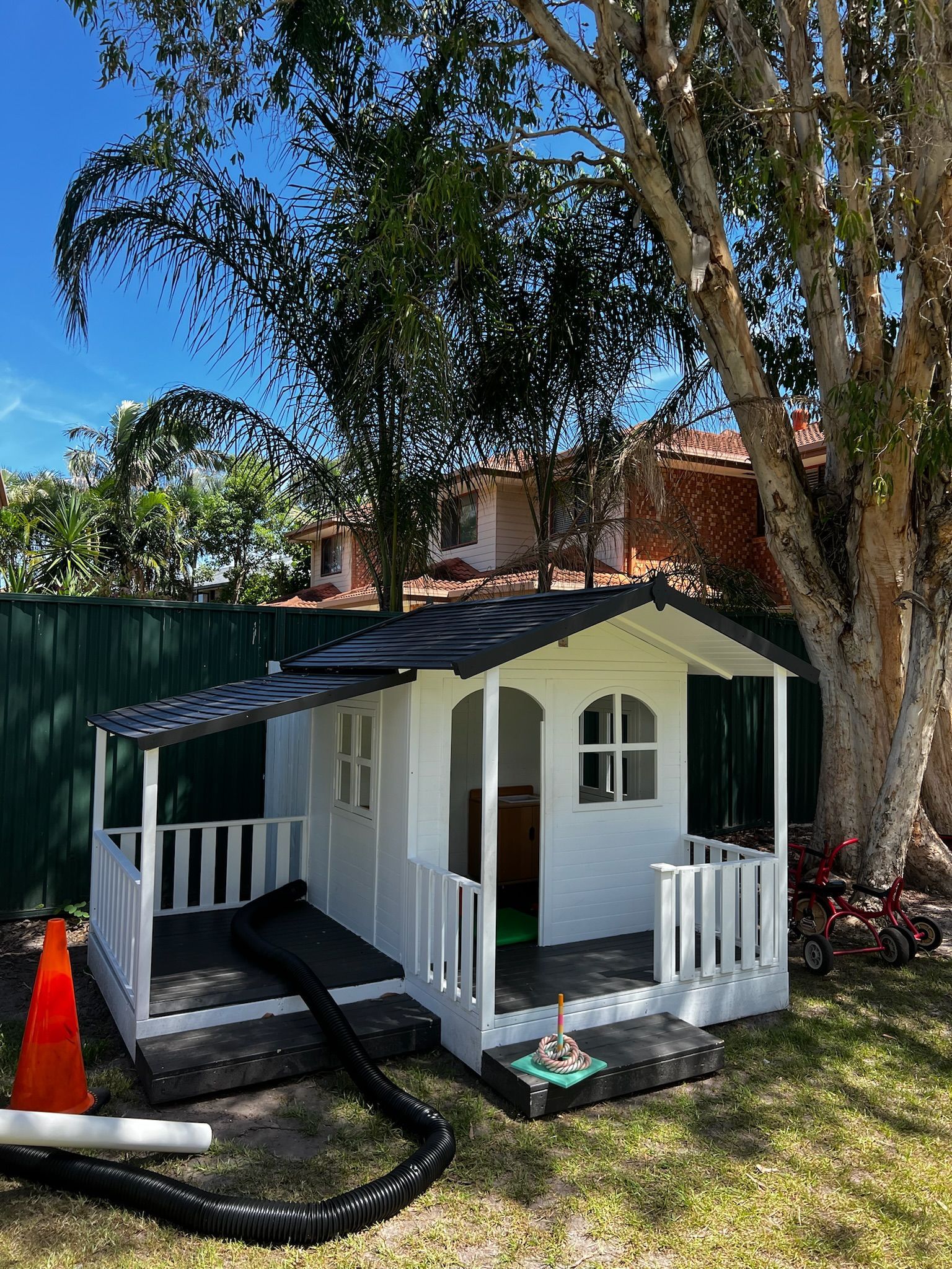 A white playhouse with a black roof and a porch in a backyard.