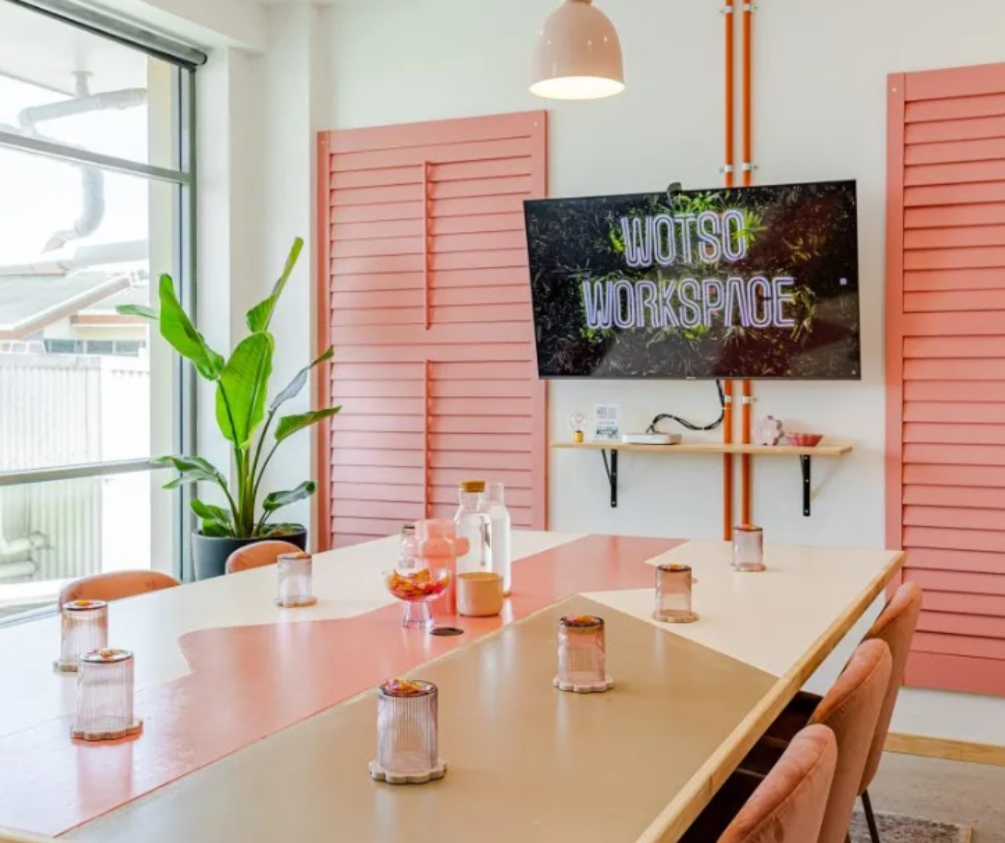 Bright meeting room with a long wooden table, pink walls, plants, and a TV reading “CO-WORKING WORKSPACE” Bright meeting room with a long wooden table, pink walls, plants, and a TV reading “CO-WORKING WORKSPACE”